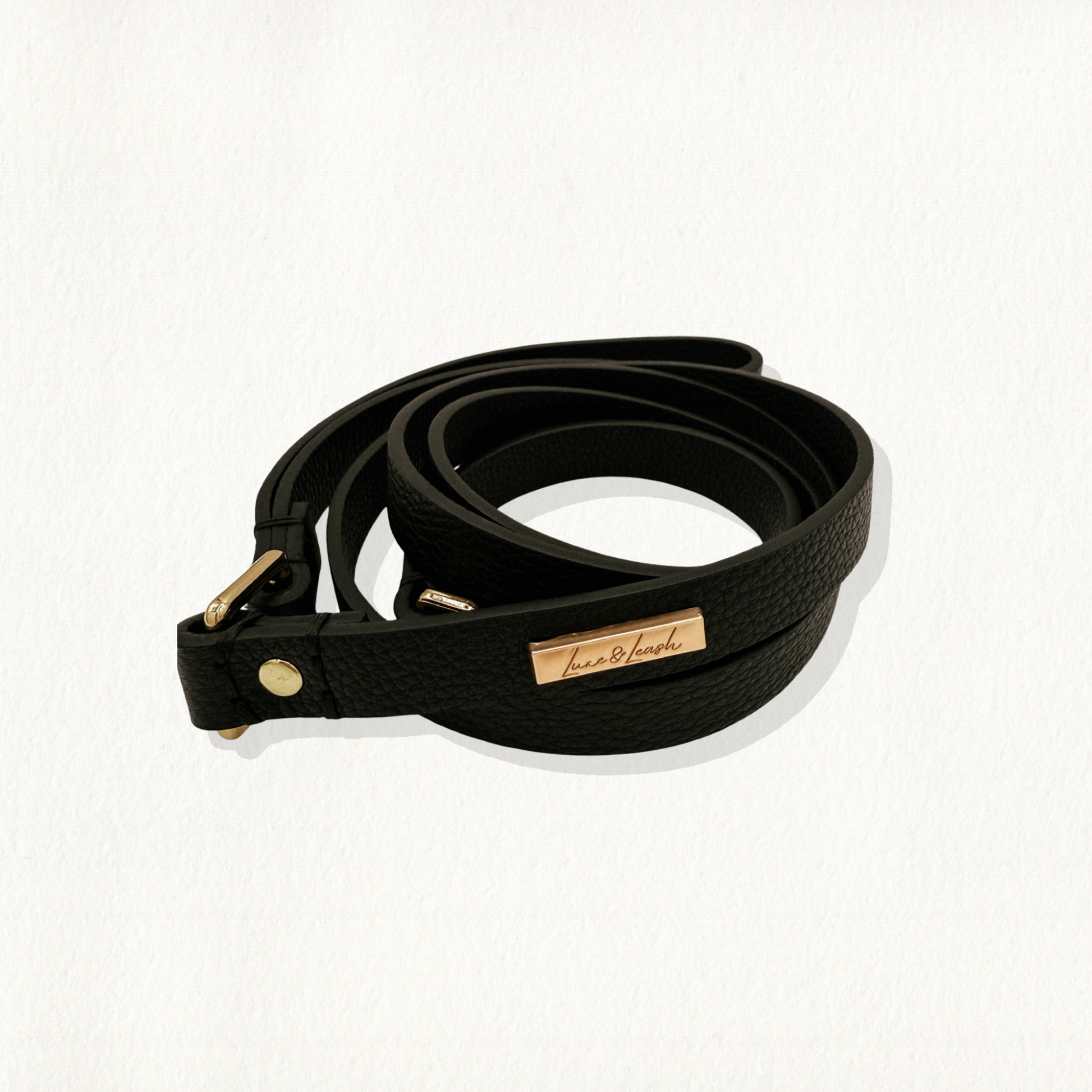 A black, pebble-textured leather dog leash with gold hardware and a "Luxe & Leash" logo plate, coiled on a white background.