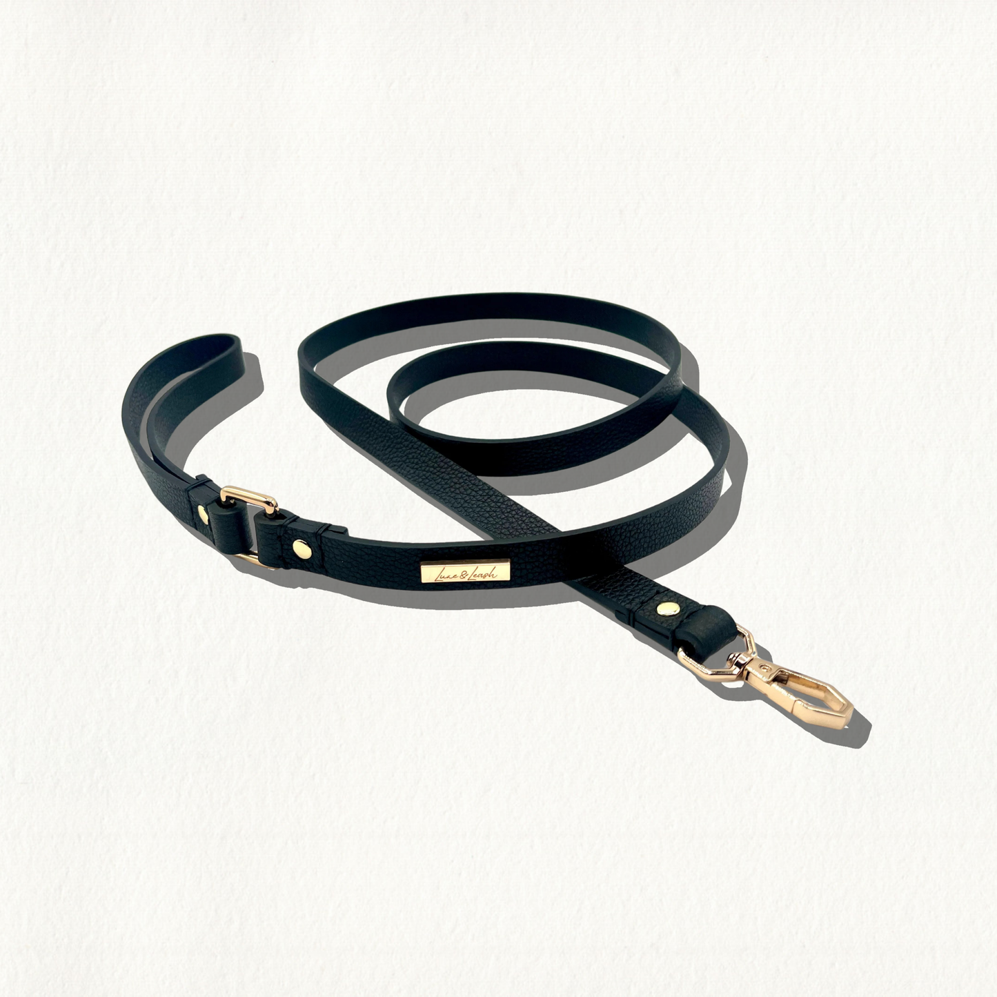 A black, pebble-textured leather dog leash with gold hardware and a "Luxe & Leash" logo plate, coiled on a white background.