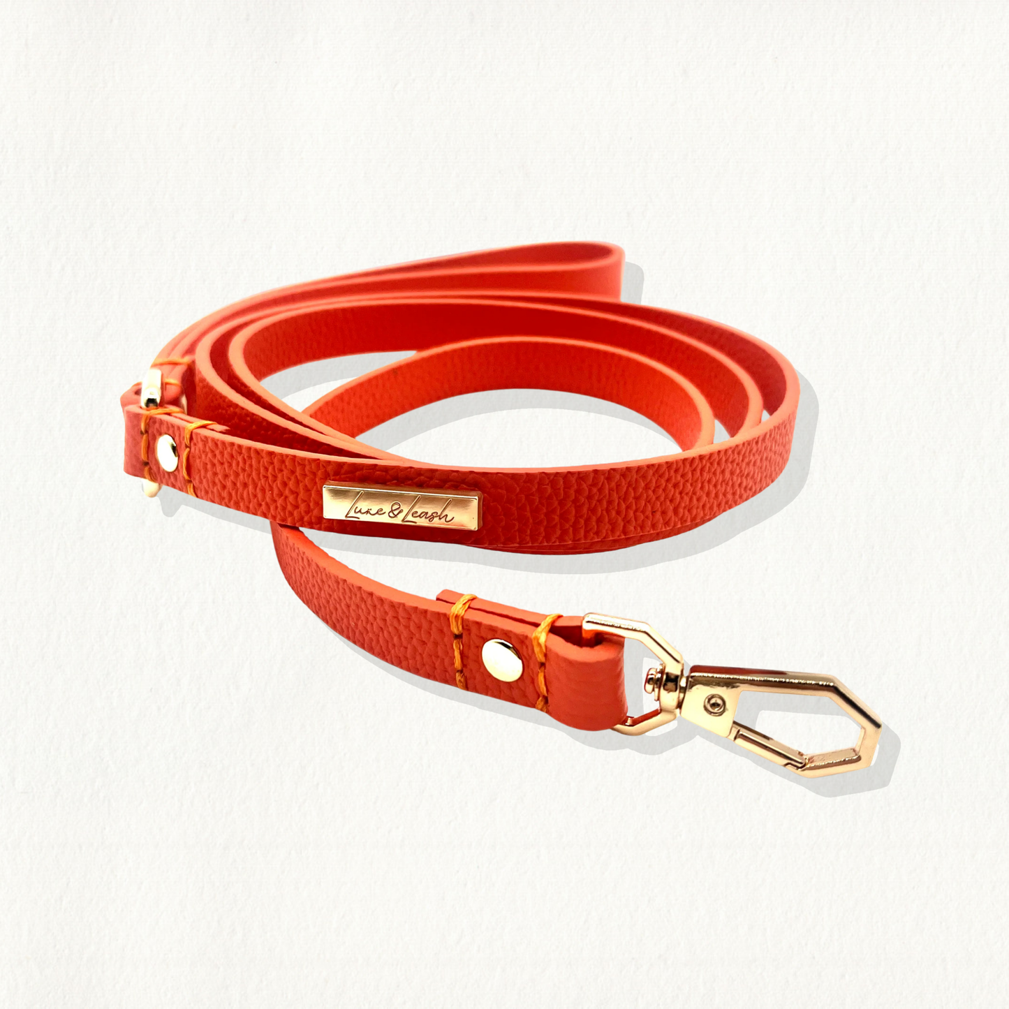 A vibrant orange, pebble-textured leather dog leash with gold hardware and a "Luxe & Leash" logo plate, coiled on a white background.