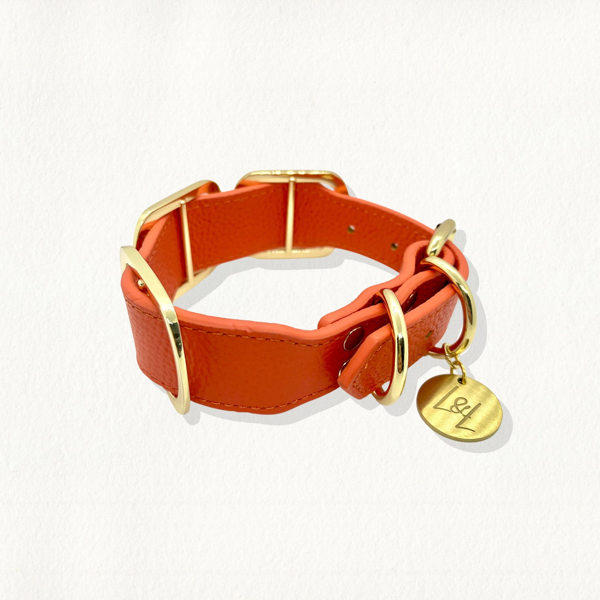 A side view of an orange luxury dog collar showing gold rectangular accents along the band and a hanging "L&L" branded gold tag.